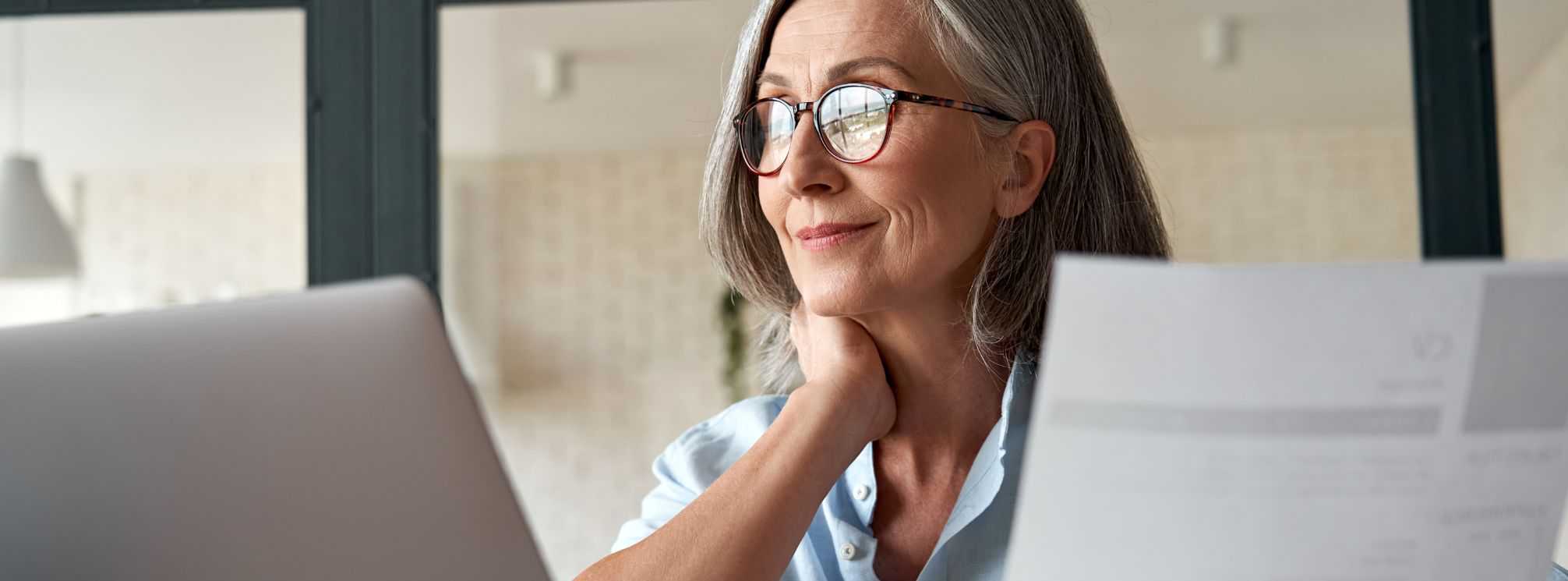 remote job interview. Older woman, wearing glasses, looking over a resume and on her laptop conducting a job interview remotely.