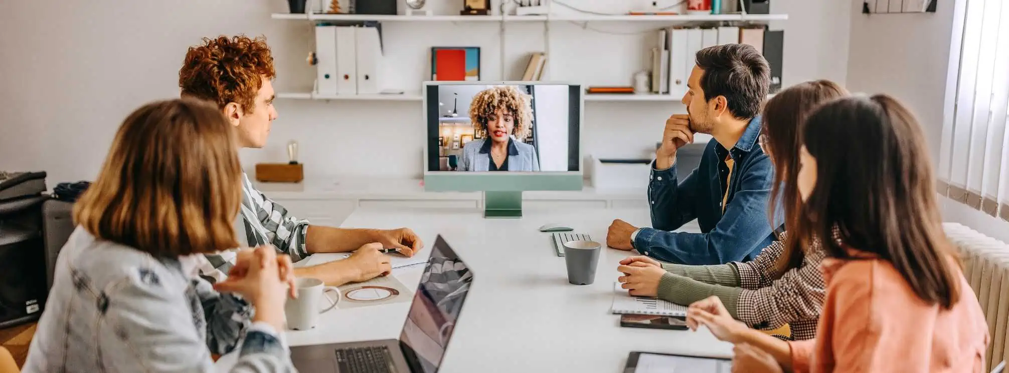 example of the hybrid workforce model in action. Five in office staff collaborating with a remote worker on a computer monitor.