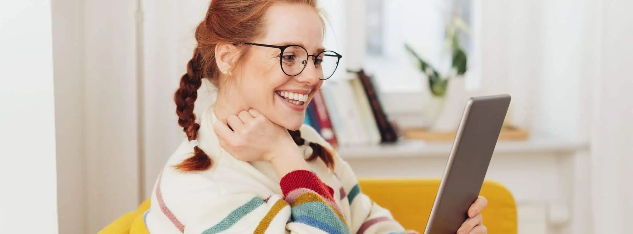 Red-haired woman in glasses, sitting in a chair in her home office and reading her employee newsletter on a tablet.