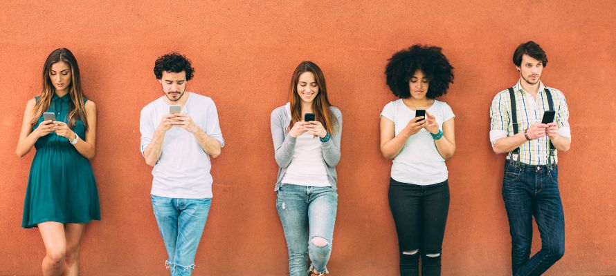 Group of five people leaning against an orange wall and texting. This is to represent sending mass texts to employees.