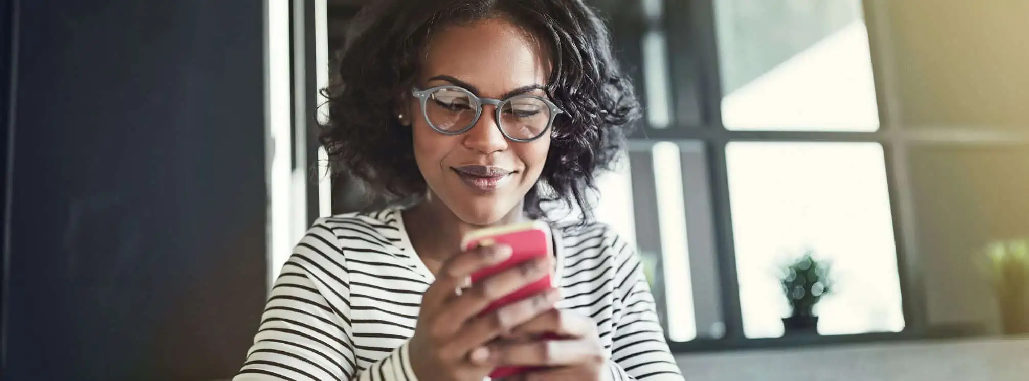 Black woman wearing glasses and a striped shirt, checking her phone from the office to see internal comms text messages