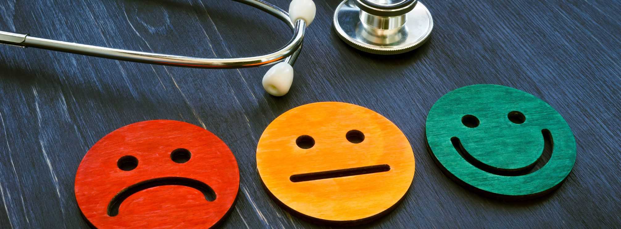 Spectrum of patient experience represented by three wooden smiley faces, one a red frown, the middle a yellow neutral face, and the right one a green smiley face. A stethoscope is on the same table by the smiley faces.