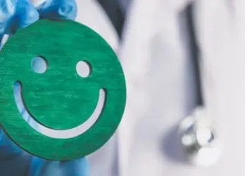 A doctor in a coat with blue gloves on holding up a green, wooden carving of a smiley face to represent a positive patient experience.