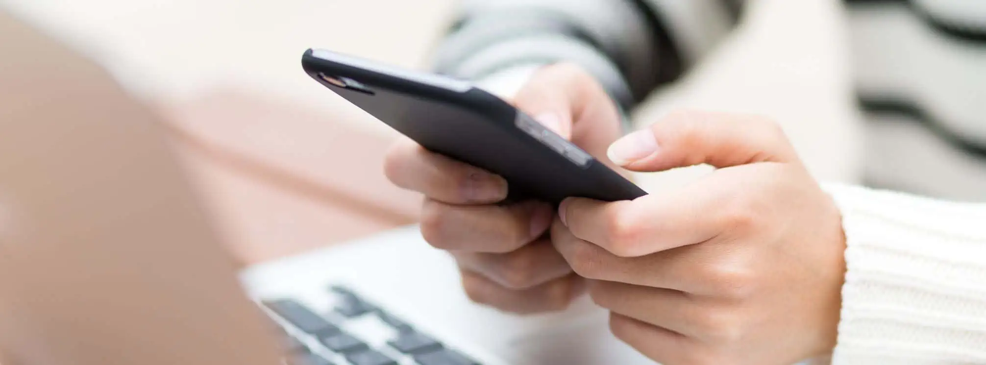 Woman's hands texting on her smartphone with her laptop open as well.