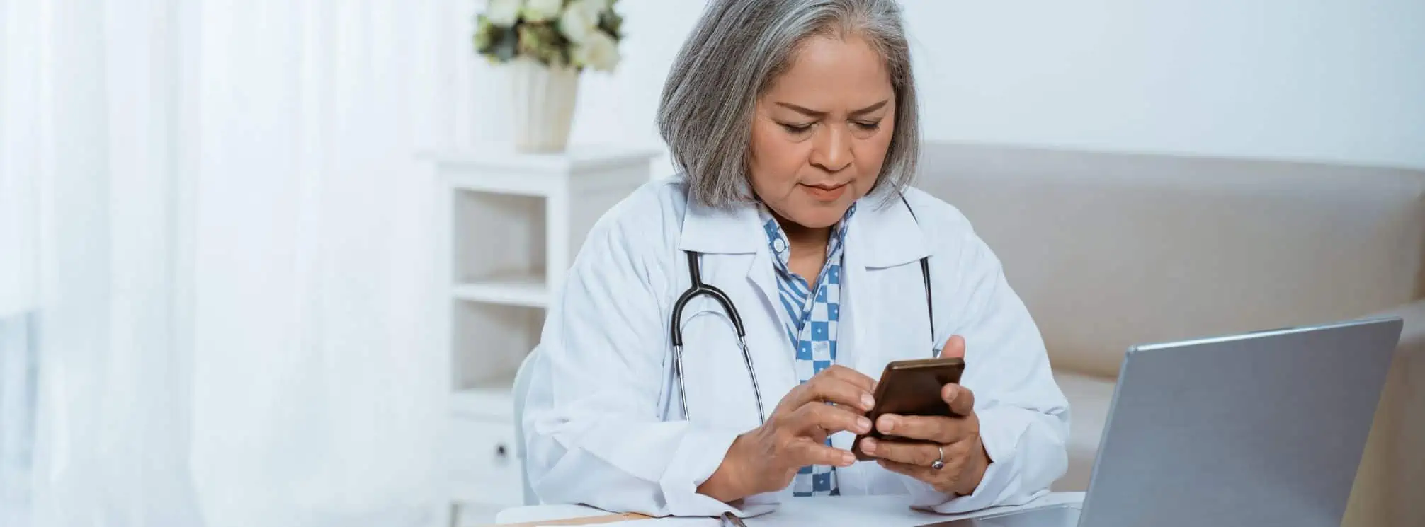 An older, asian doctor checking her smartphone while working on her laptop in a break room.