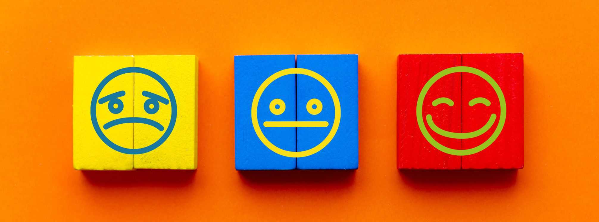 Vibrant orange background with three wooden blocks on it. One is yellow with a frowning face drawn on it, the middle block is blue with a neutral facial expression, and the last is red with a smiley face on it.