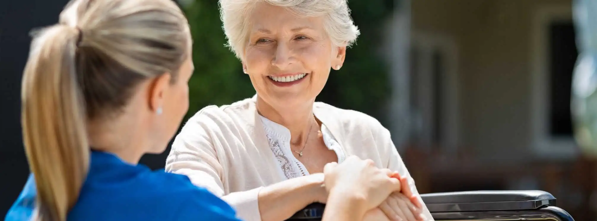 Young nurse in blue scrubs sitting with an elderly woman in a wheelchair.