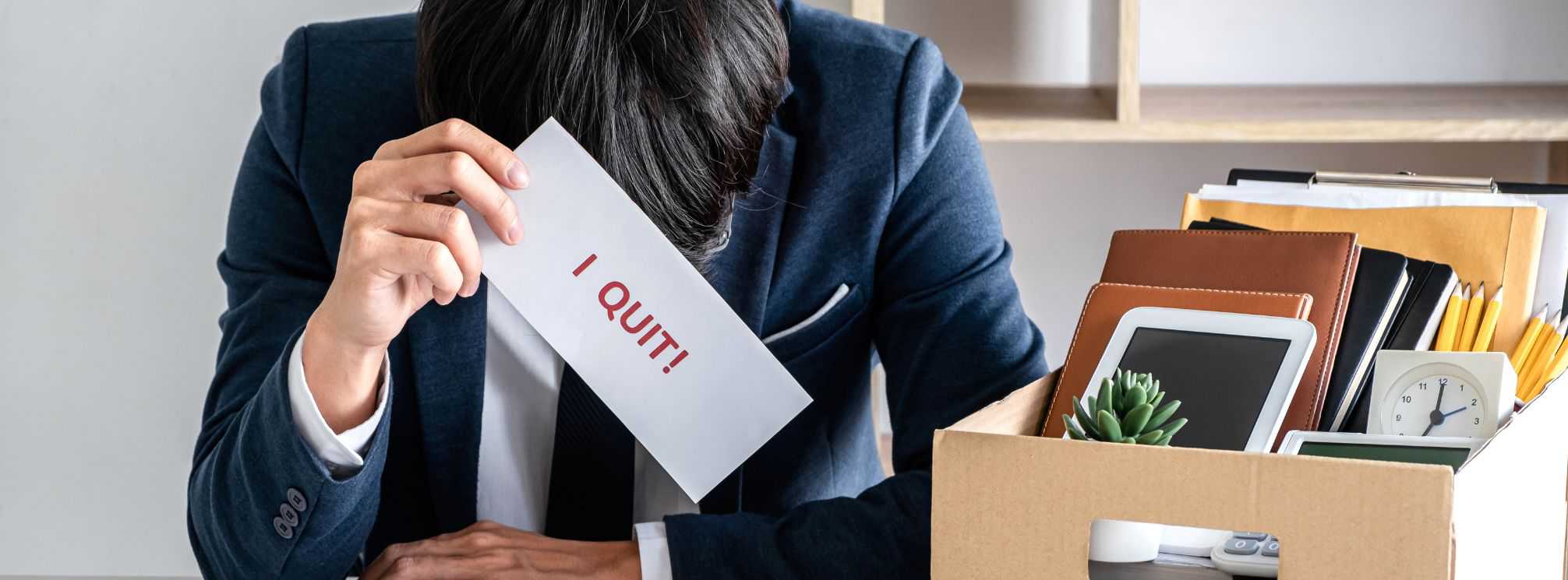 Despondent employee sitting next to a box of their office possessions, holding a sign that says "I quit!"