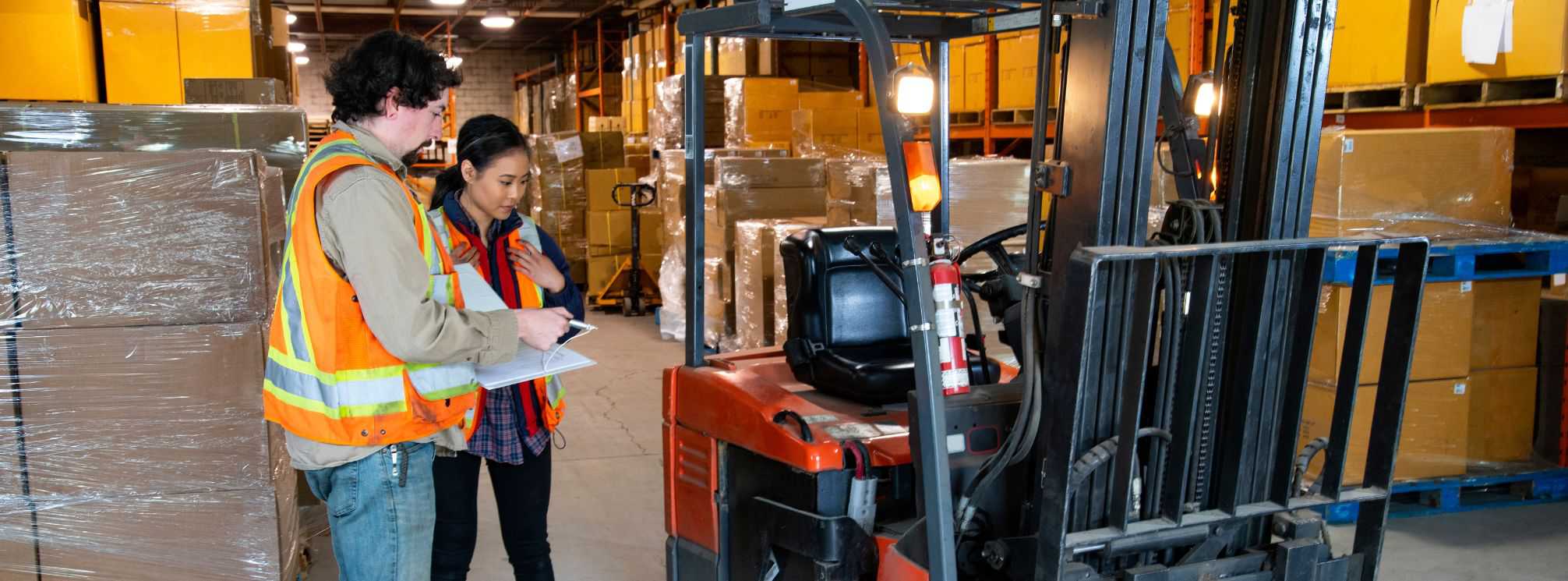 Two warehouse employees in safety vests, reviewing the steps for operating a forklift.