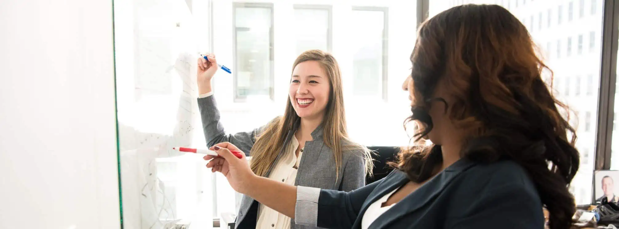 Two women working together at a whiteboard using dry erase markers and smiling.