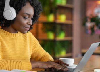 Black woman in a yellow sweater wearing over-ear headphones and going through an employee learning and development course on her laptop.